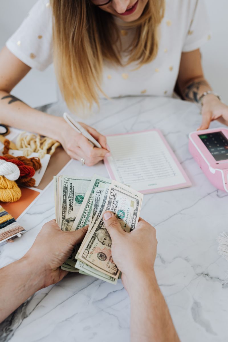 Two friends reviewing a shared expense list on a phone while sitting at a kitchen table.